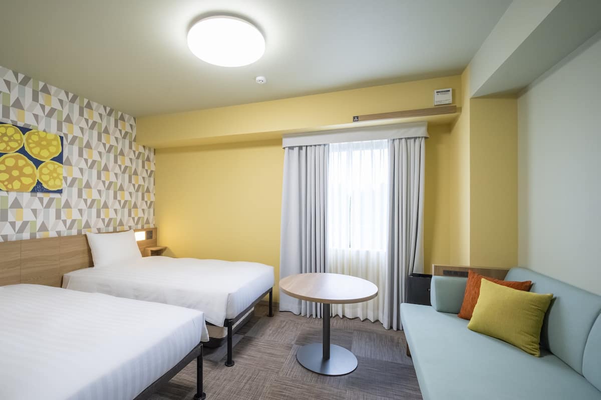 Habitación de hotel con dos camas, una mesa redonda, un sofá, pared con diseño y una ventana con cortinas.