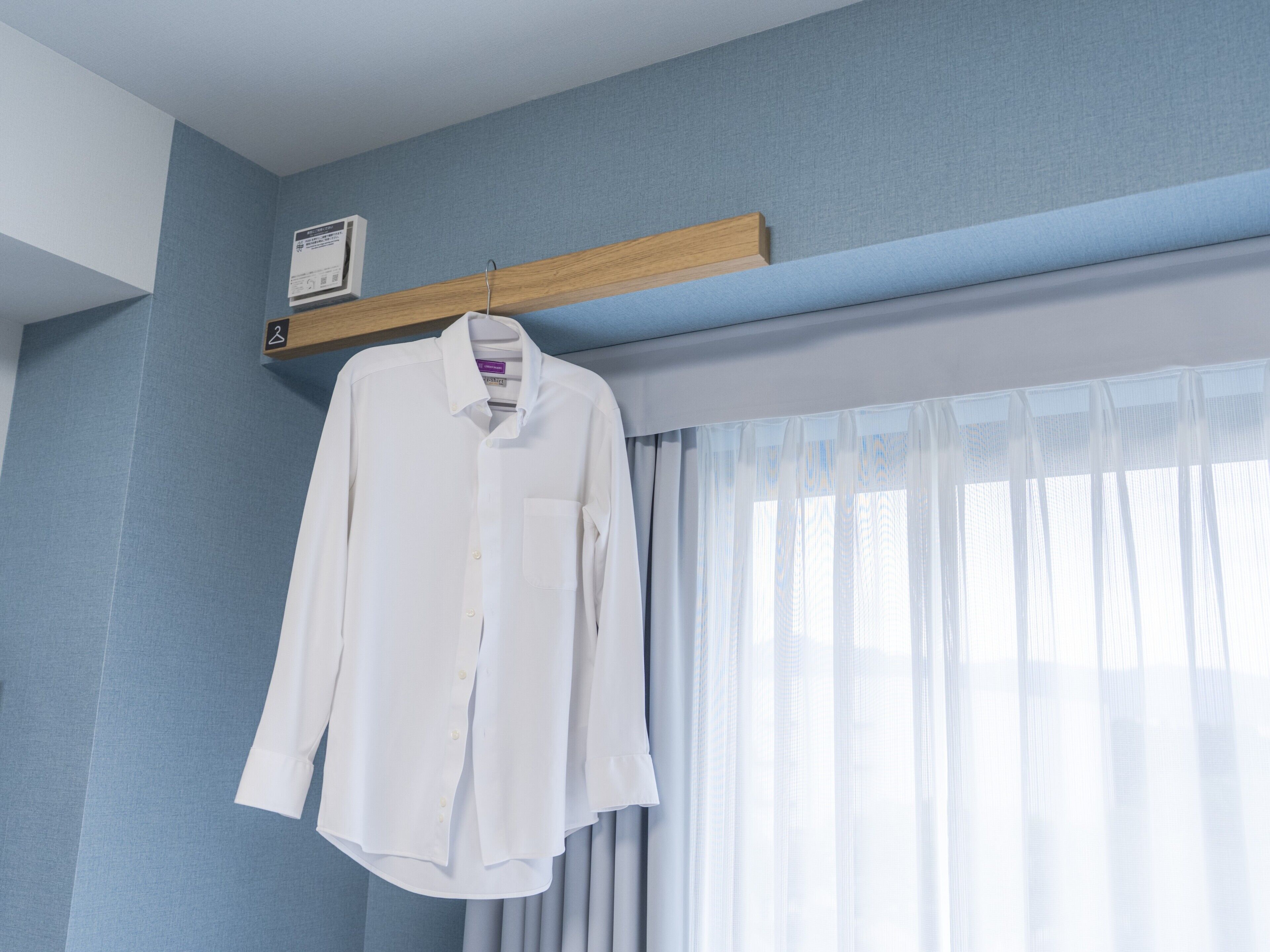 A white shirt hanging on a wooden hanger in a room with light blue walls, a window with sheer curtains, and a small wall-mounted device.
