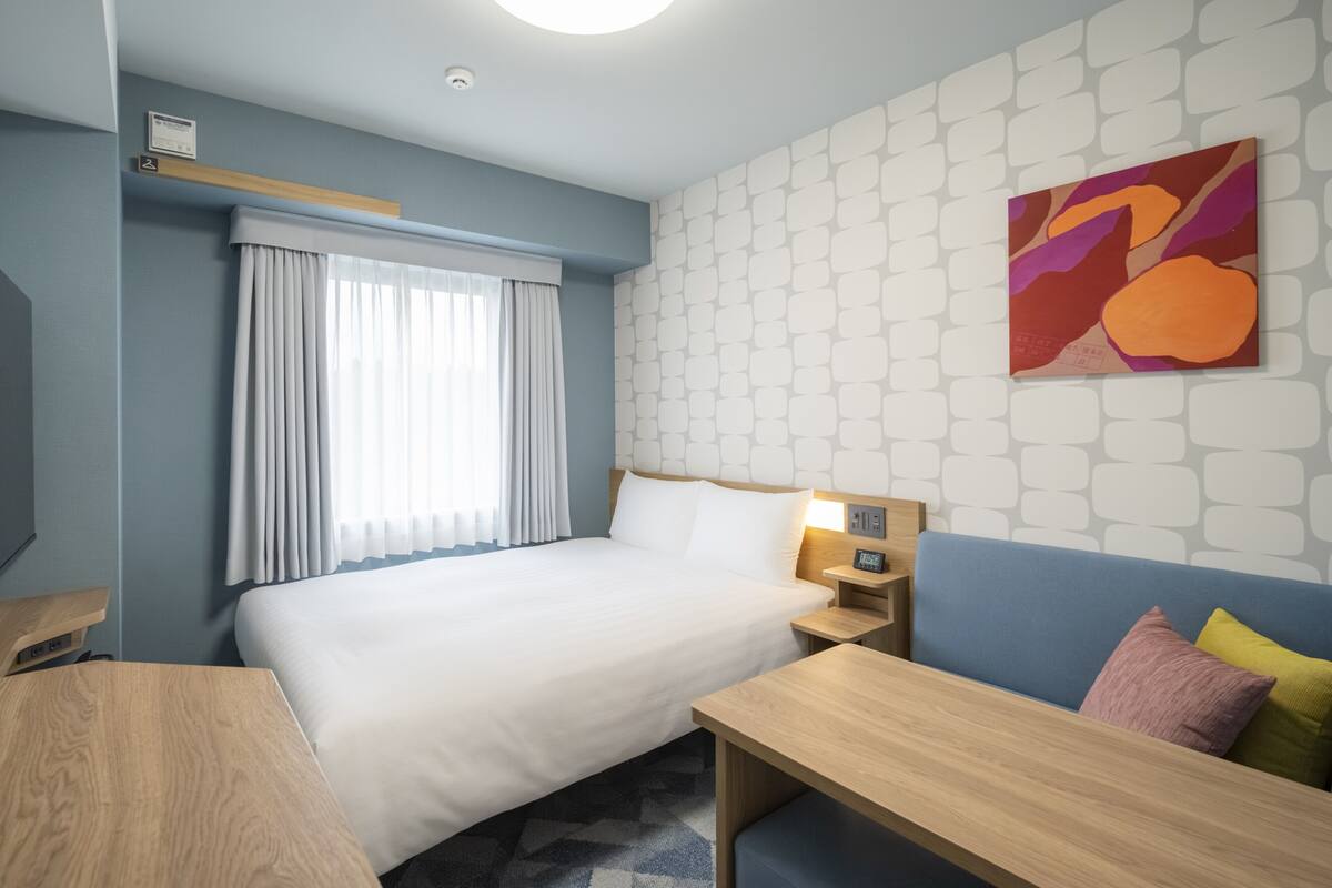 Habitación de hotel con cama, mesita de noche, lámpara y un cuadro abstracto colorido en la pared.