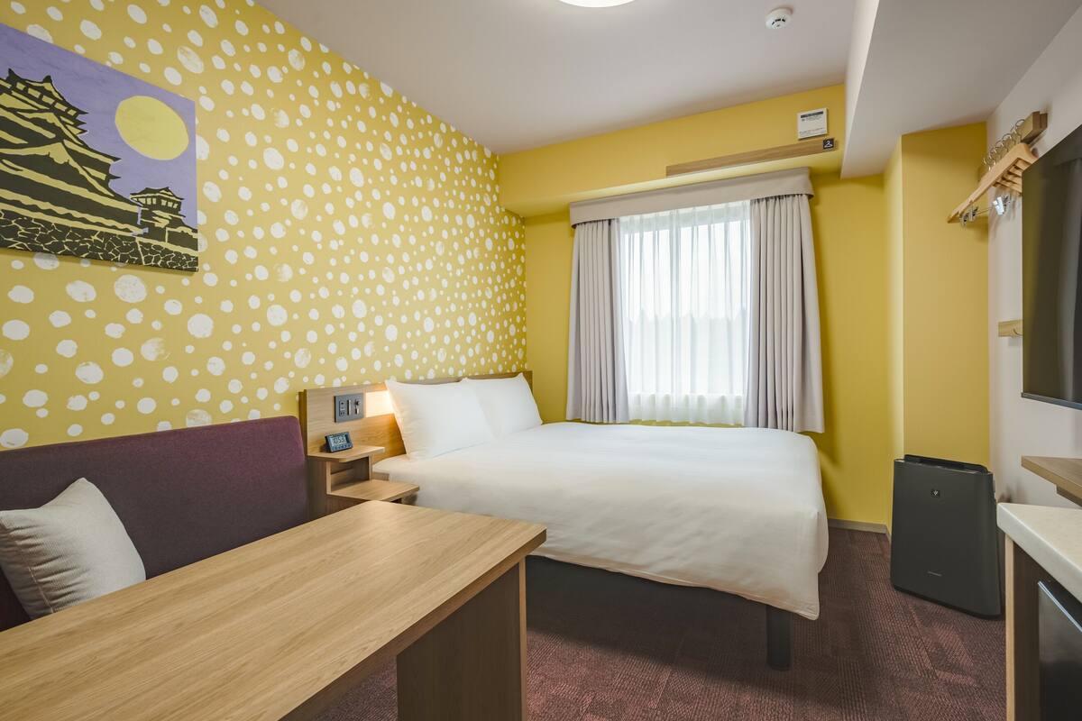 Habitación de hotel con cama, escritorio, televisión, ventana con cortinas, reloj de pared y un cuadro.