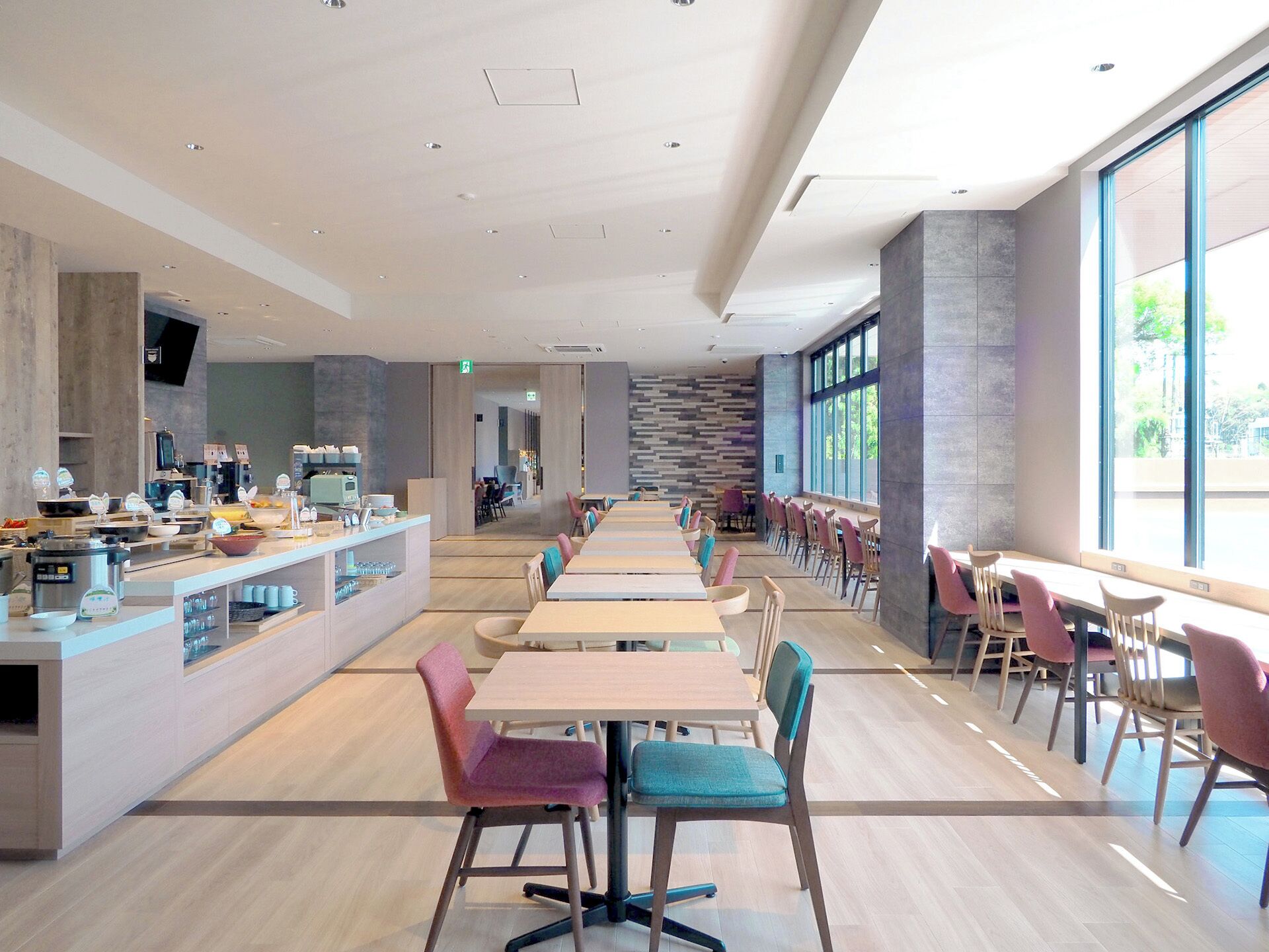 A modern dining area with long tables and chairs, a counter with food items, and large windows providing natural light.