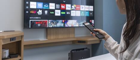 A person holding a remote control in front of a flat-screen TV displaying various streaming service logos.
