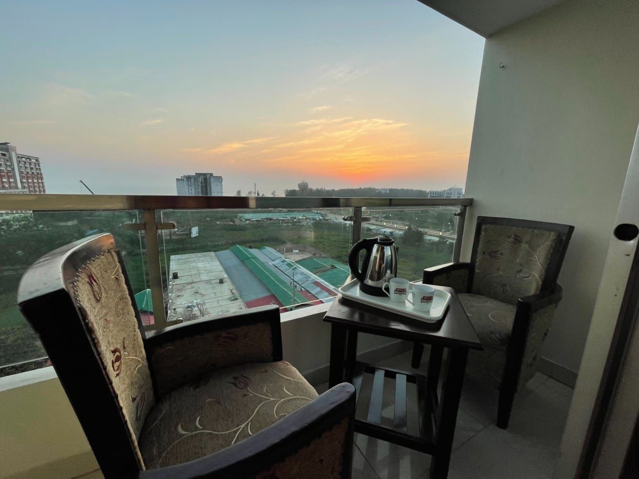 Un balcone con un tavolo, due sedie e vista su un campo sportivo al tramonto.