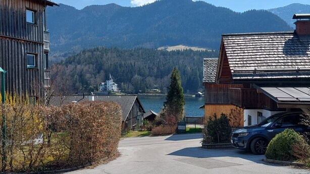 A residential area with a view of mountains, a lake, and a wooden building with a car parked in front. - Grundlsee vacation apartment - Charming vacation apartment right on the lakeshore (Mosern)