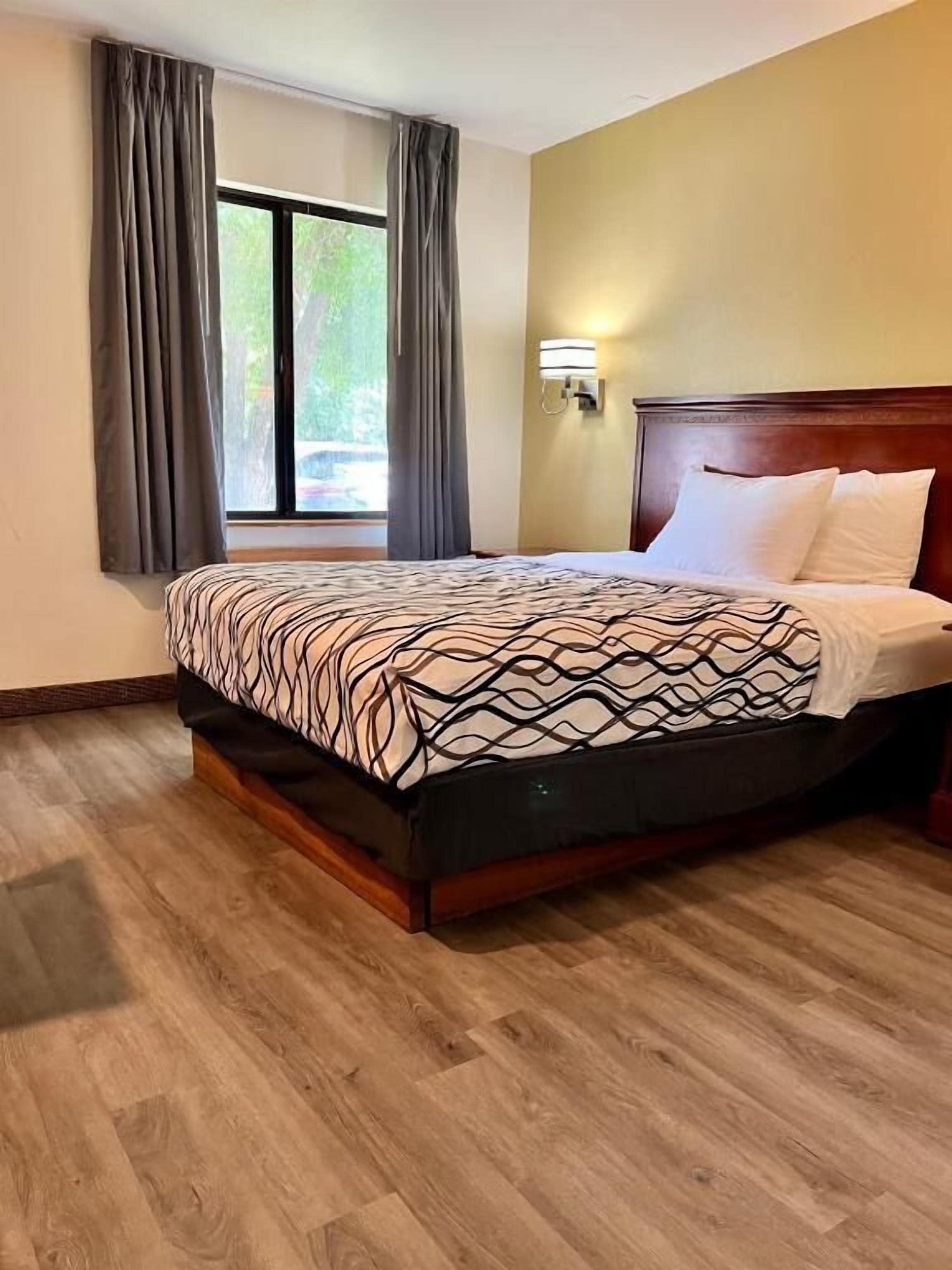 A hotel room with a bed, wooden flooring, a window with curtains, and a wall-mounted light fixture.