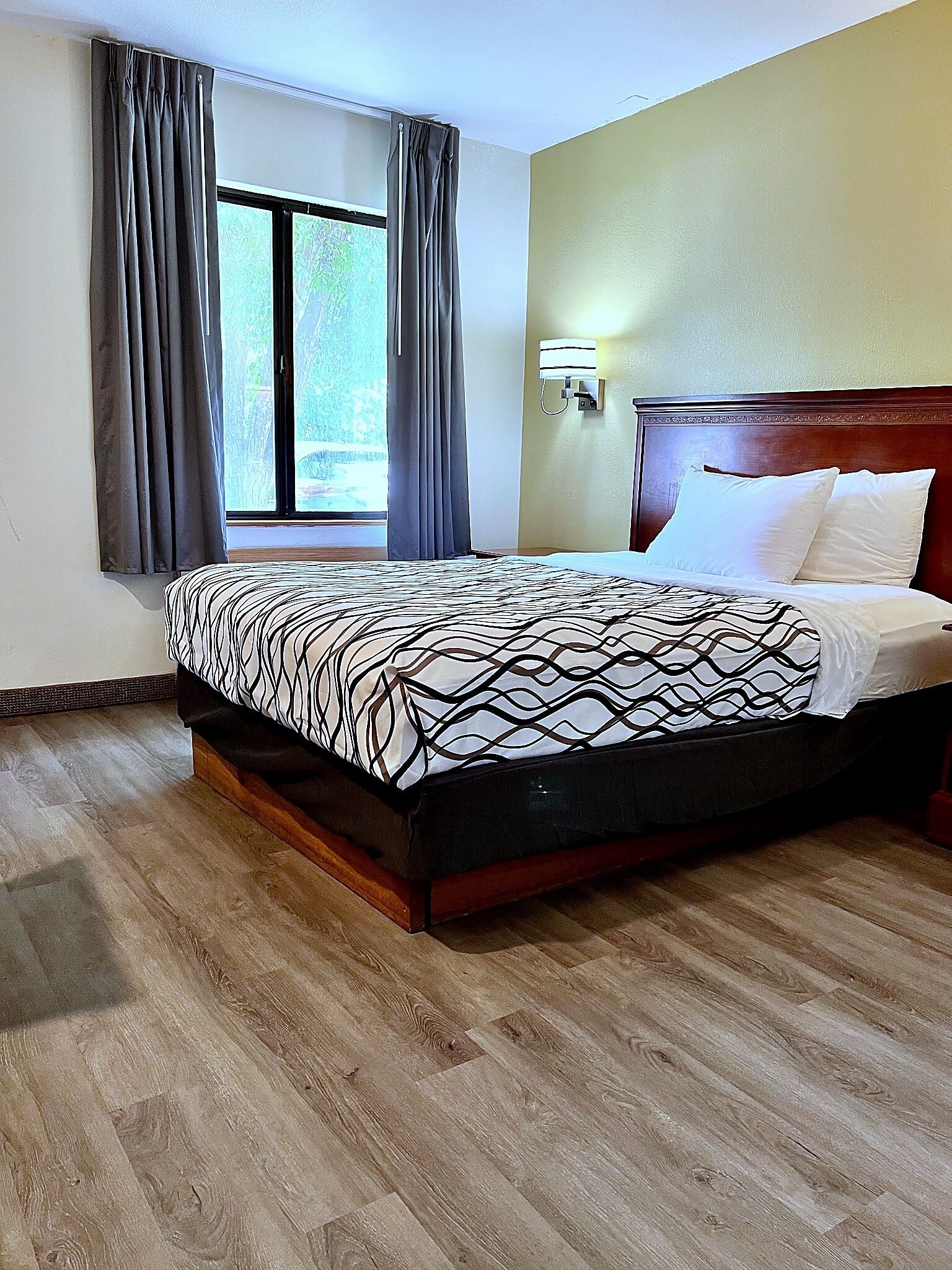 Habitación de hotel con una cama grande, suelo de madera, una ventana con cortinas y una lámpara de pared.