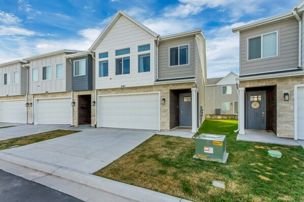 A row of modern townhouses with attached garages, a well-maintained lawn, and a utility box on the grass.
