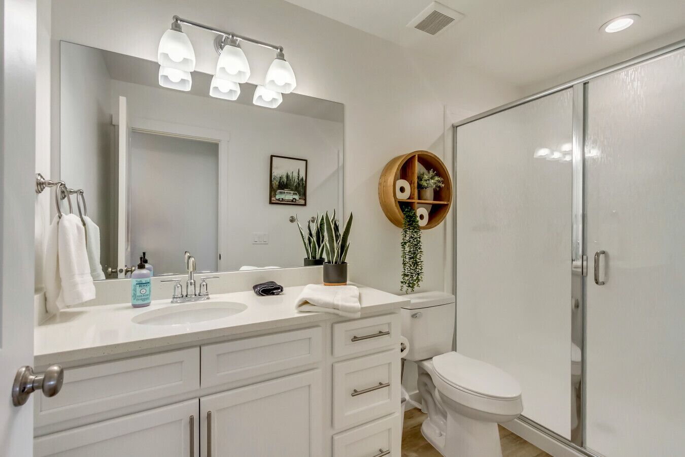 Un baño con lavamanos blanco, espejo y retrete, ducha con puerta de cristal y estante de madera con artículos de aseo.