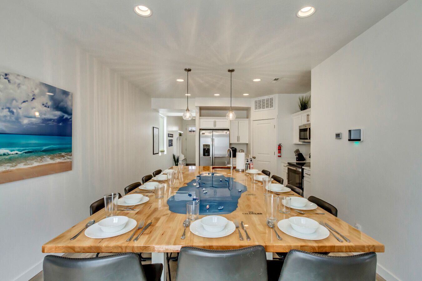 Une longue table à manger avec des chaises, un coin cuisine et un tableau représentant une scène de plage.