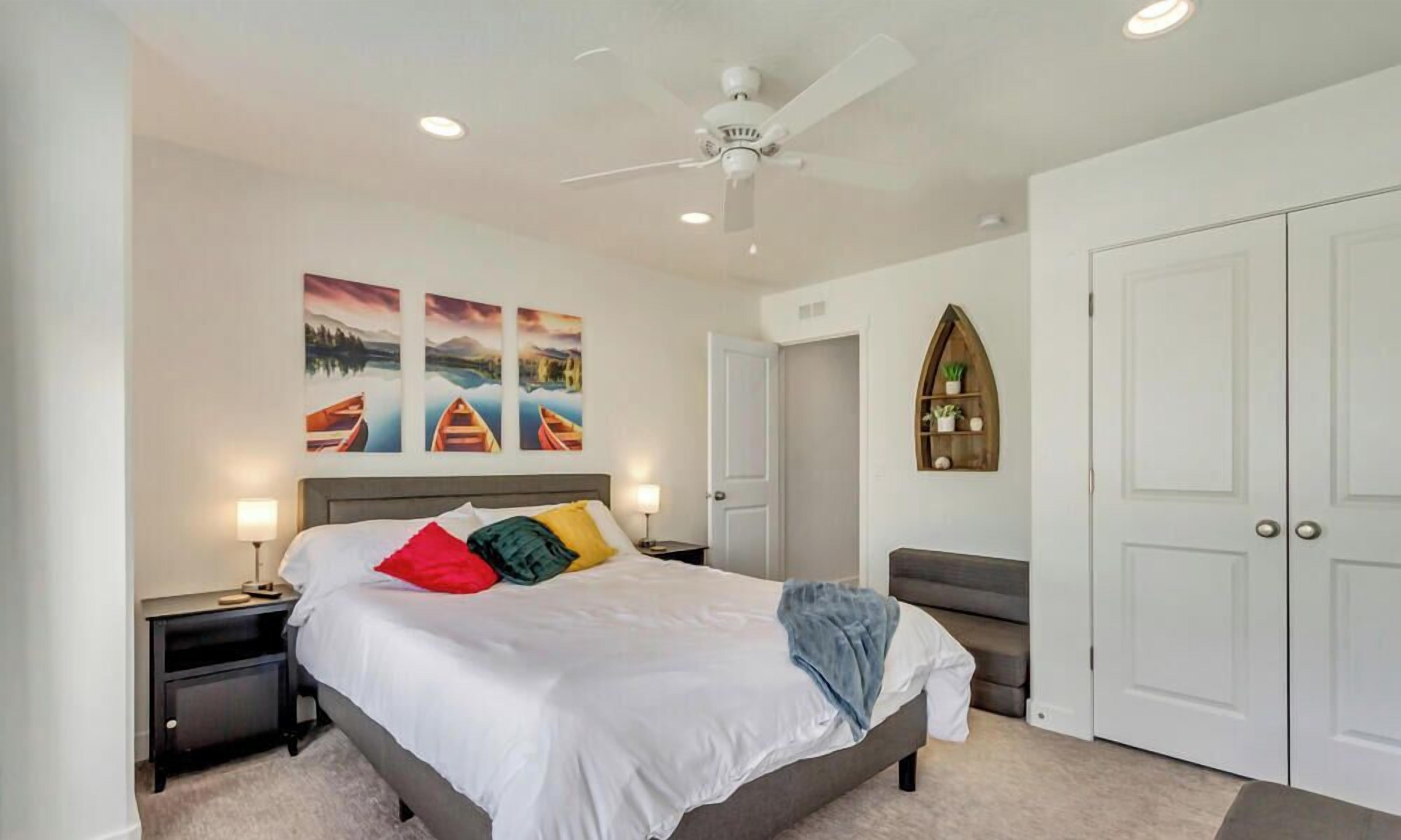 Ein Schlafzimmer mit einem Bett, einem Deckenventilator, einem Nachttisch, einem Schminktisch, einer Kommode, einer Bank, einer Tür und einem wandmontierten Regal.