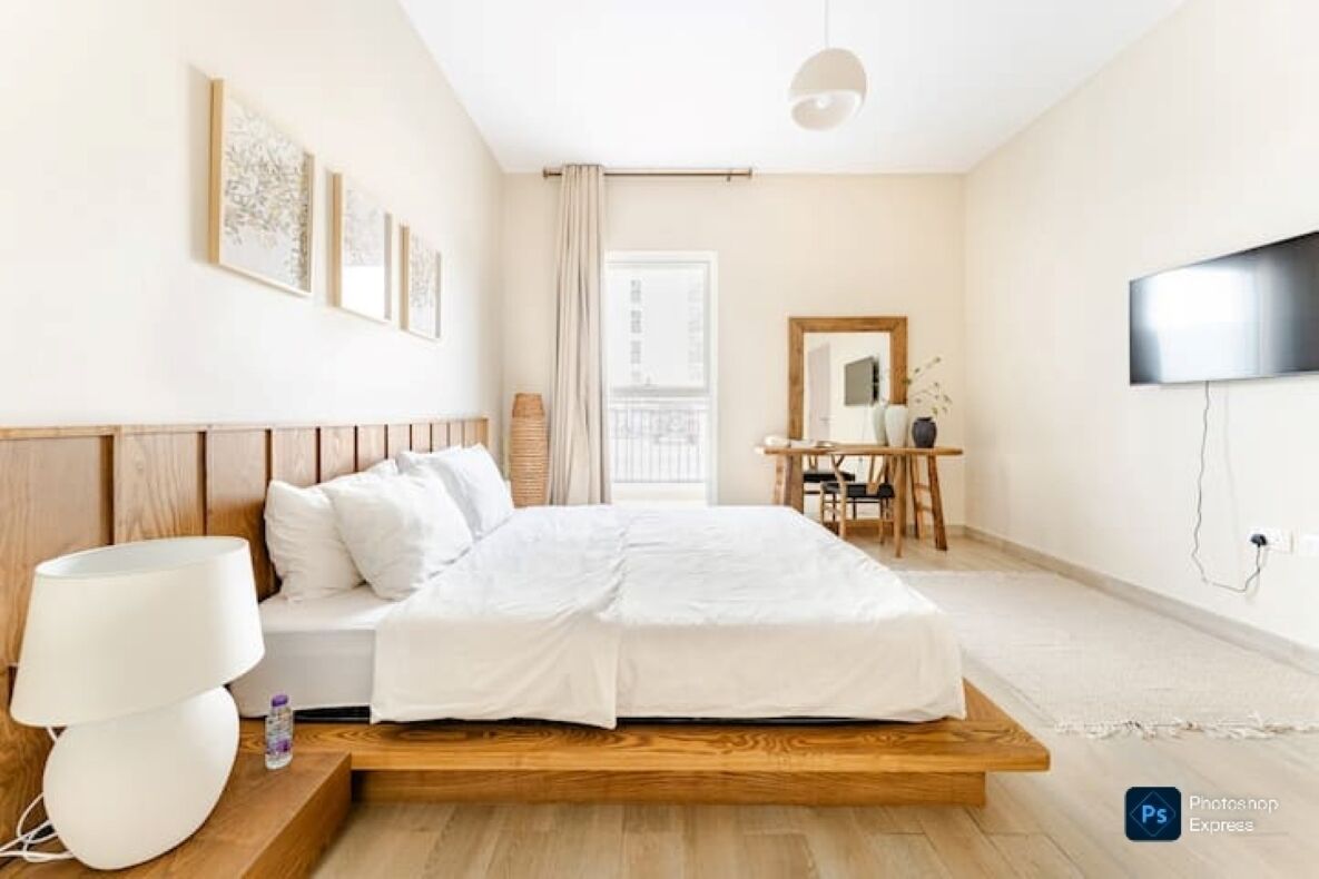 A modern hotel room with a wooden bed frame, white bedding, a desk with a chair, a flat-screen TV, and framed artwork on the wall.