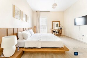 Ein modernes Hotelzimmer mit einem Bettgestell aus Holz, weißer Bettwäsche, einem Schreibtisch mit Stuhl, einem Flachbildfernseher und gerahmten Kunstwerken an der Wand.