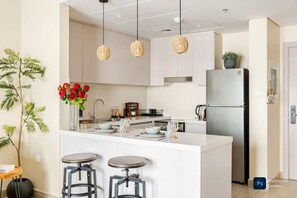 Eine moderne Küche mit weißen Schränken, einer zentralen Kochinsel mit Sitzgelegenheiten, hängenden Pendelleuchten und einem Kühlschrank.