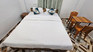 Un lit bien fait, avec une literie blanche, deux coussins décoratifs, une table de chevet en bois, une petite table avec une chaise et des carreaux de sol à motifs.
