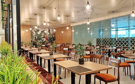 A modern restaurant with wooden tables, orange cushioned chairs, hanging pendant lights, and a large mural of parrots.