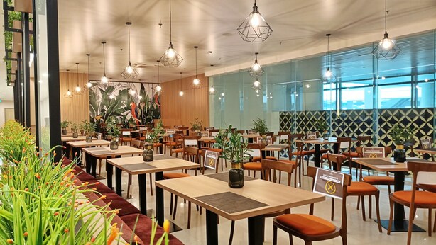 A modern restaurant with wooden tables, orange cushioned chairs, hanging pendant lights, and a large mural of parrots. - Cordia Hotel Yogyakarta  (Temon)
