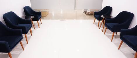 Una habitación con piso blanco, pared blanca y un armario de madera. Seis sillas azul oscuro con patas de madera dispuestas en dos filas.