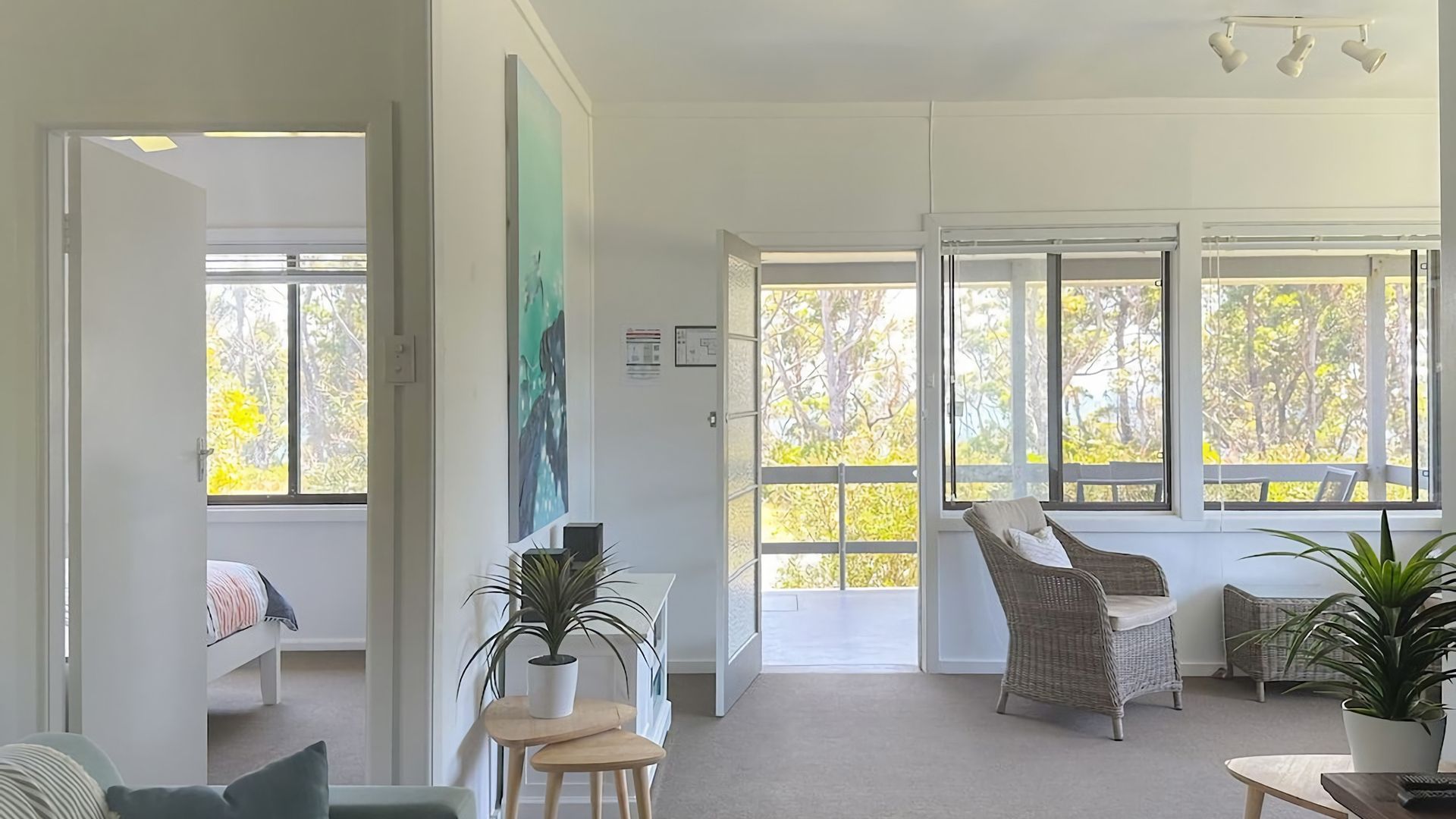 Una habitación luminosa con ventanas grandes, una silla de mimbre, una mesita y una cama.