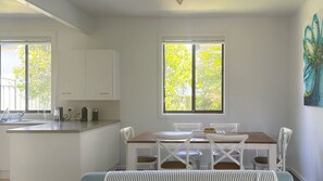A modern kitchen with white cabinetry, a dining area with a wooden table and white chairs, and a large window with a view of greenery. - Yonder is near the ocean and beach (Bendalong)