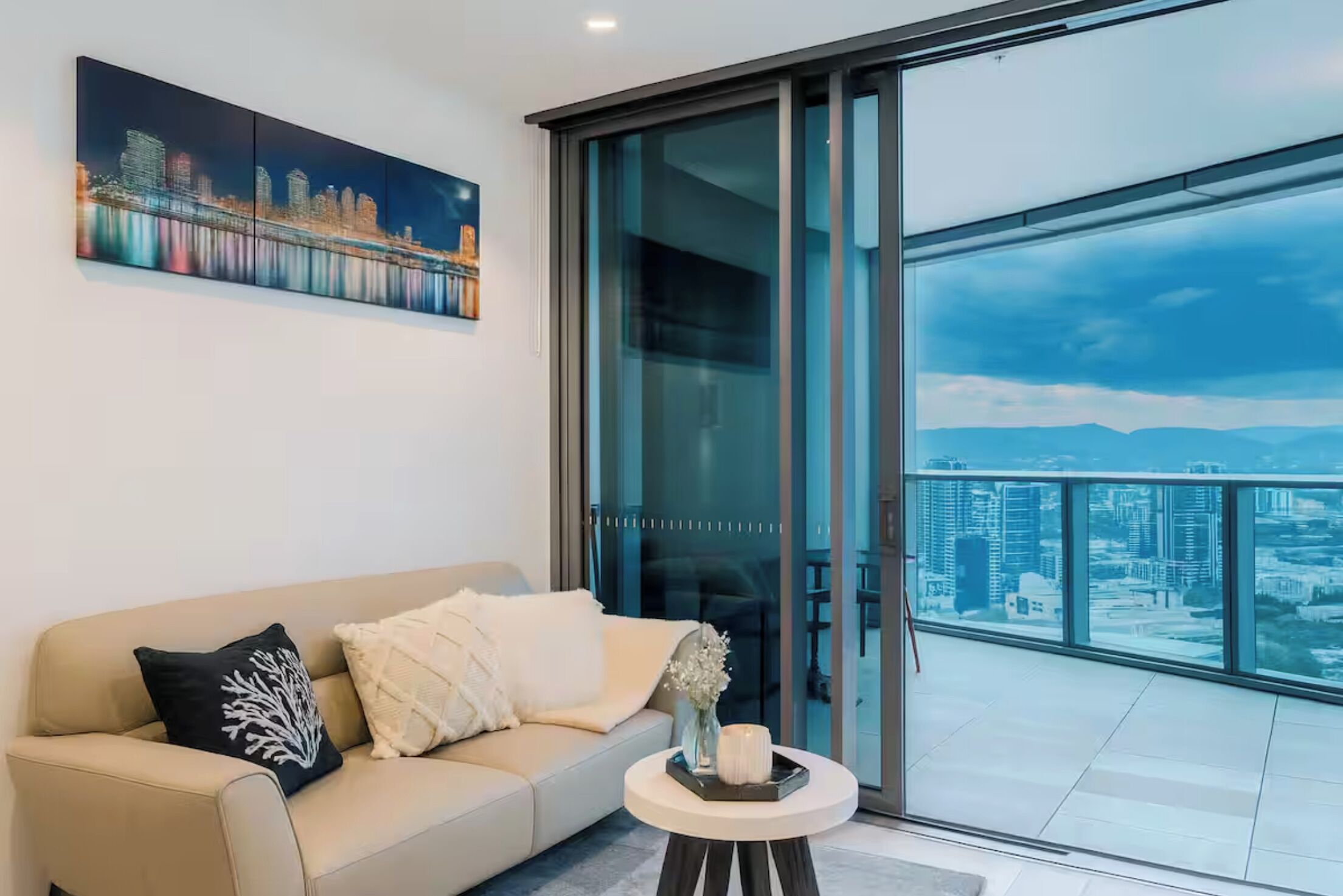A modern living room with a sofa, a glass door leading to a balcony with a city view.