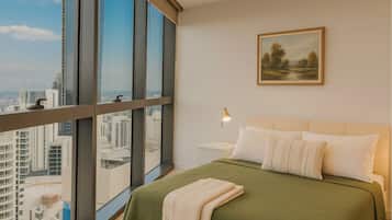A bedroom with a large window, a bed with a green bedspread, white pillows, a bedside lamp, and a framed landscape painting on the wall.