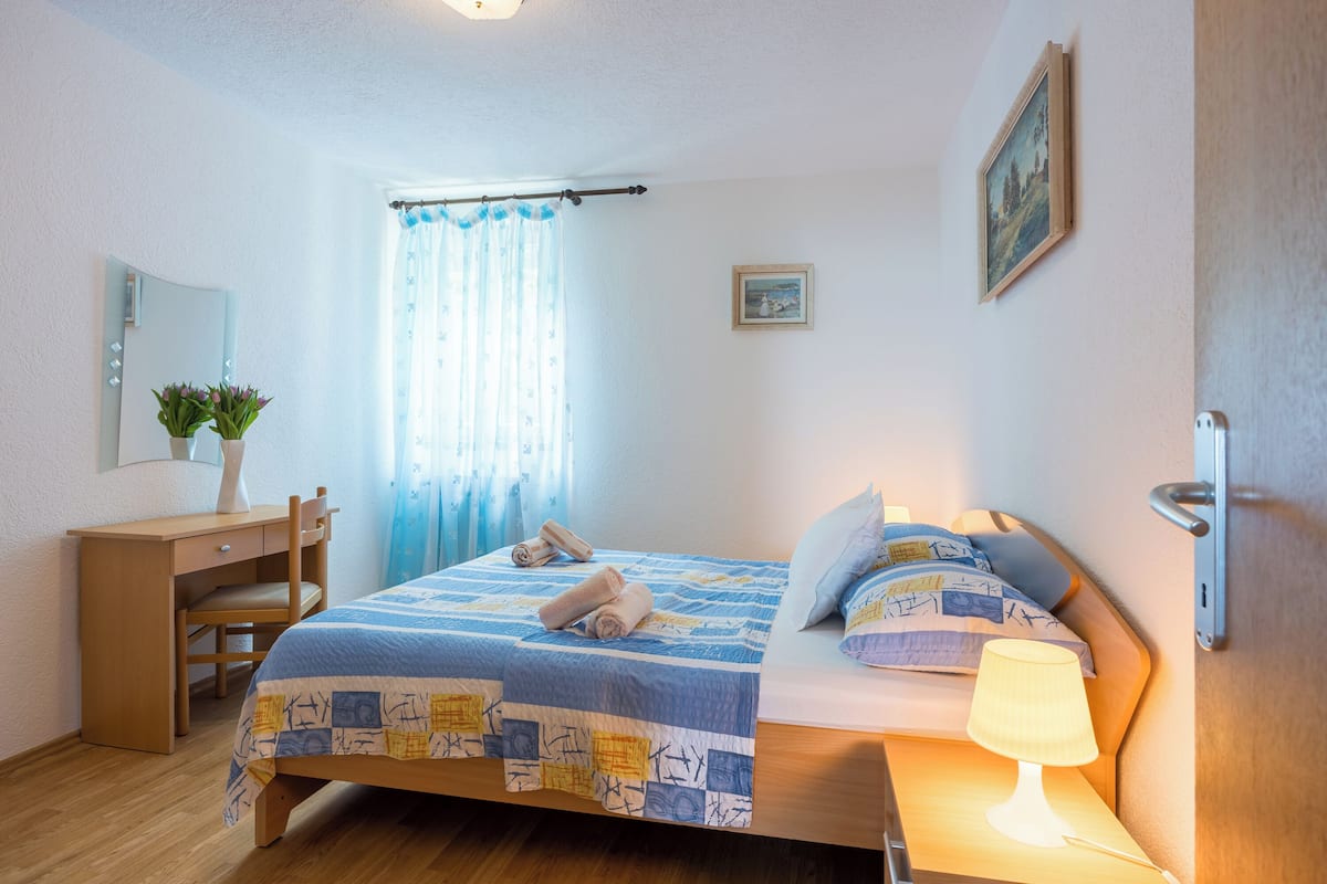 Une chambre à coucher comprenant un lit, une table de chevet, une lampe, un bureau, une chaise et une fenêtre avec des rideaux.