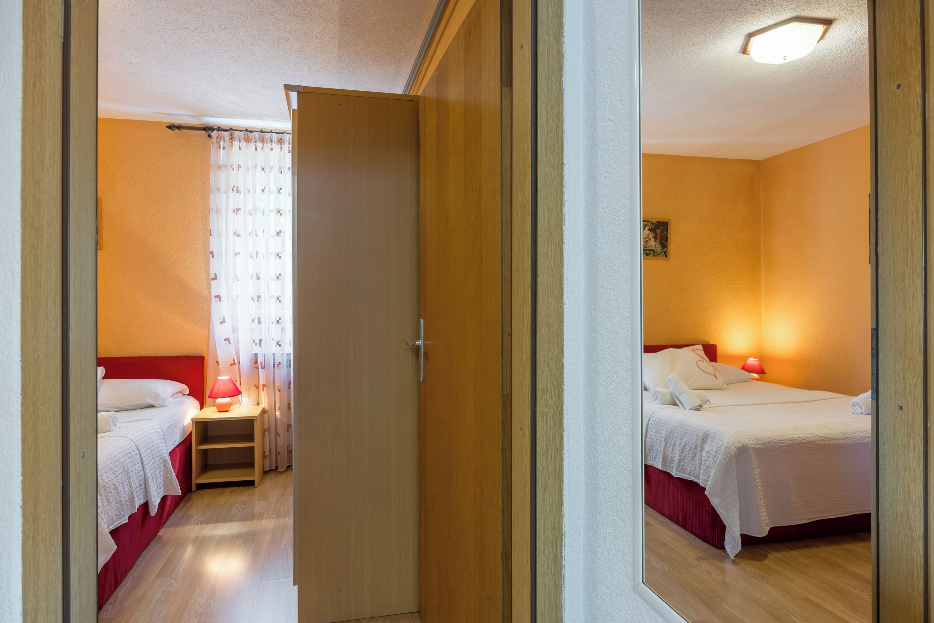 Habitación de hotel con dos camas, una mesita de noche con lámpara, y una puerta que da a otra habitación.