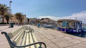 A waterfront promenade with a bench, boats docked, and a building with signage for tours. - Amalija sea View Apartments (Mali Losinj)