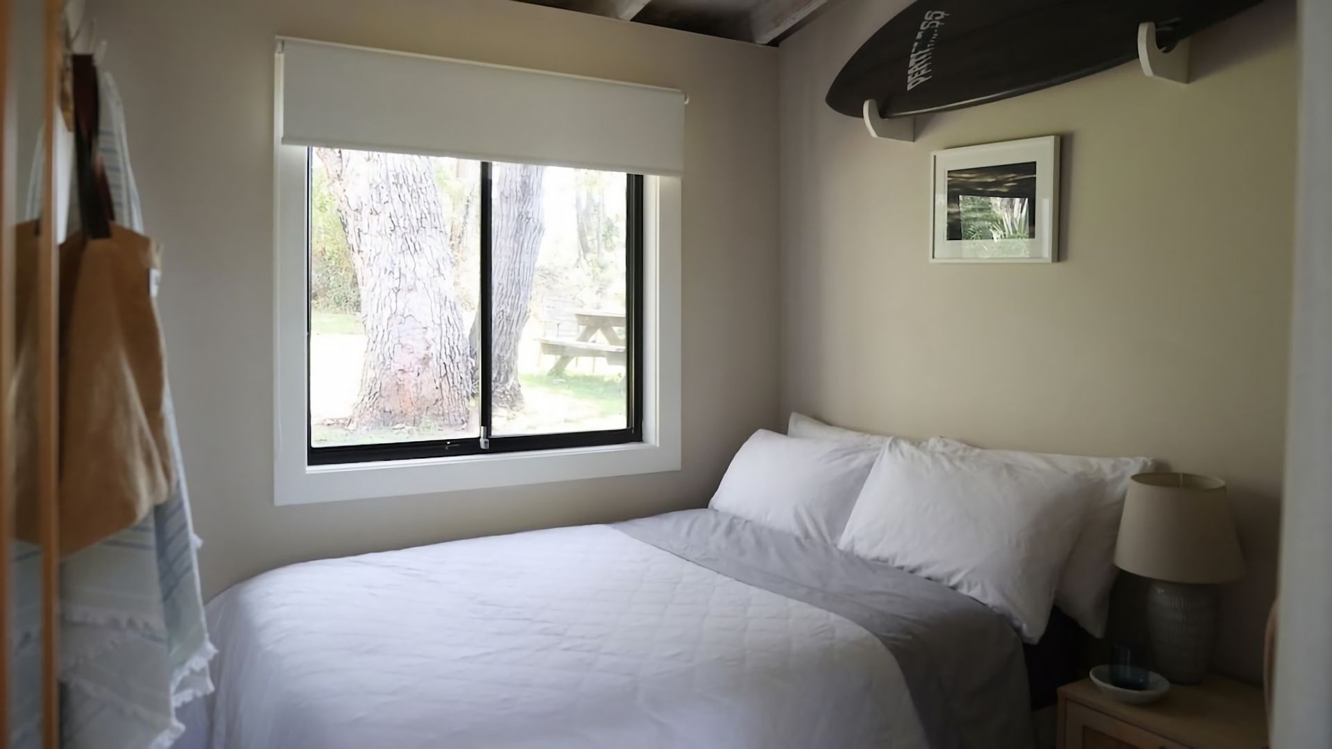 A bedroom with a bed, a window, a bedside table with a lamp, and a framed picture on the wall.