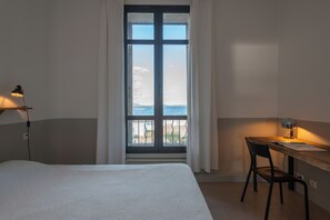 A hotel room with a bed, a desk with a chair, a desk lamp, and a view of the sea through a large window. - magnificent 2 bedrooms with sea and salt marsh views (Porto-Vecchio)