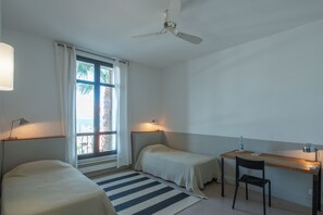 Ein Zimmer mit zwei Einzelbetten, einem Schreibtisch, einem Stuhl, einem Deckenventilator und einem Fenster mit Vorhängen.