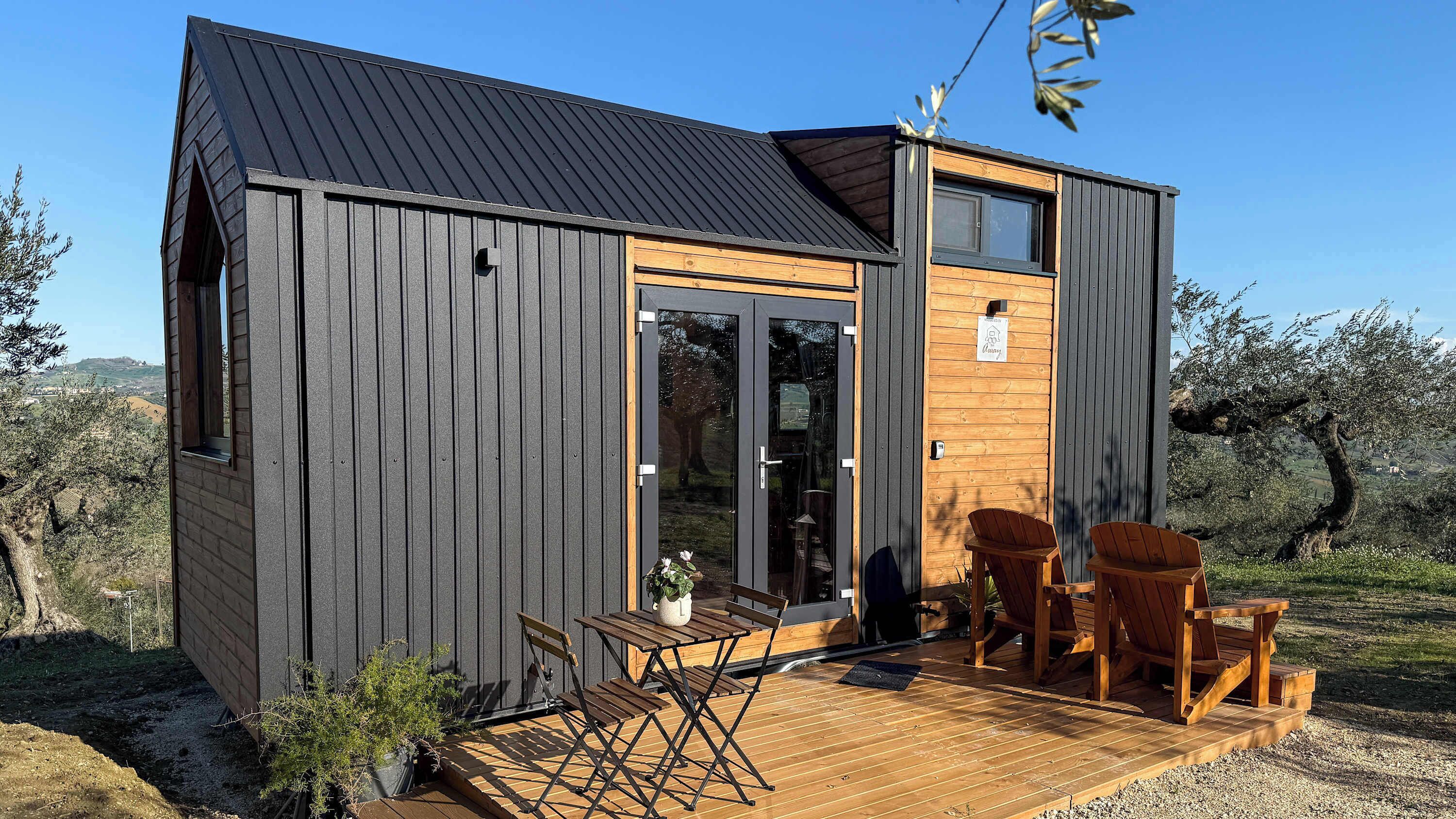 A compact cabin with a dark exterior, a wooden door, and a small deck with chairs and a table.