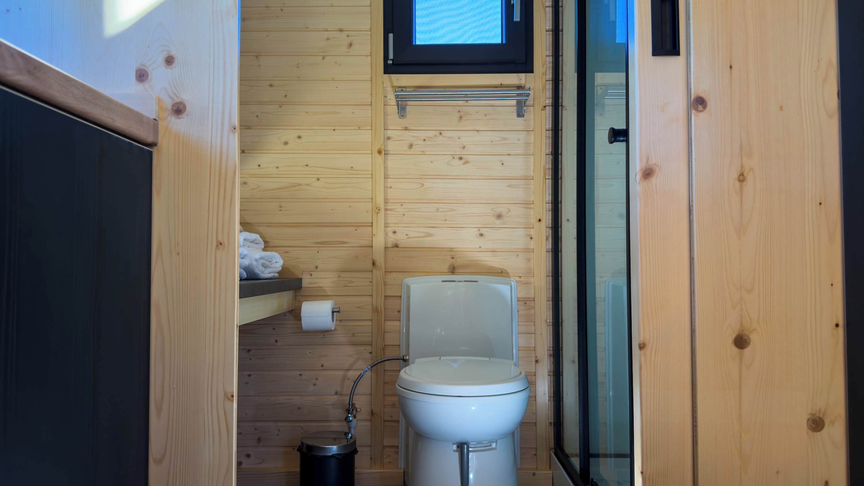 A compact wooden bathroom with a toilet, a small shelf with towels, and a window above the toilet.