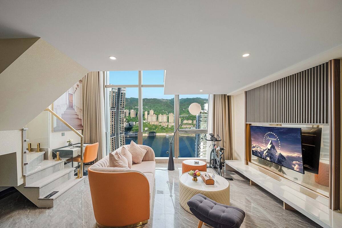 Een moderne woonkamer met een grote televisie, een bank, een salontafel, en een trap naar een buitenbalkon met uitzicht op een stadsbeeld en water.