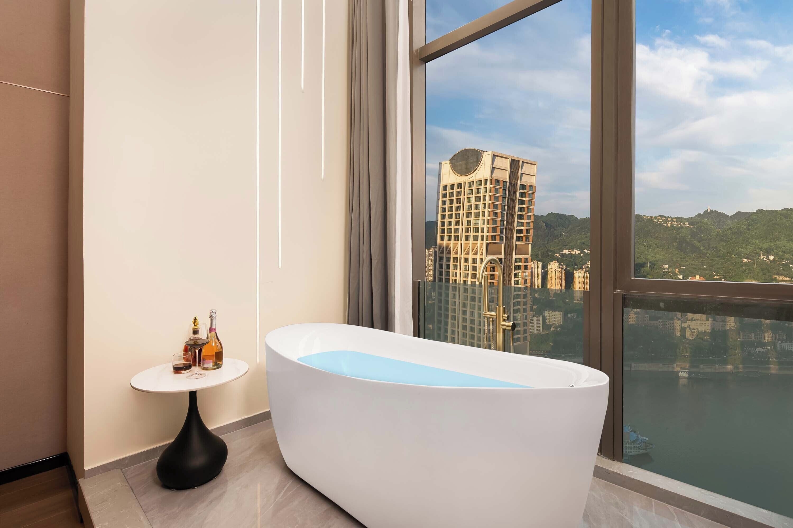 Un baño moderno con una bañera independiente, una pequeña mesa redonda con botellas y un amplio ventanal con vistas a la ciudad.
