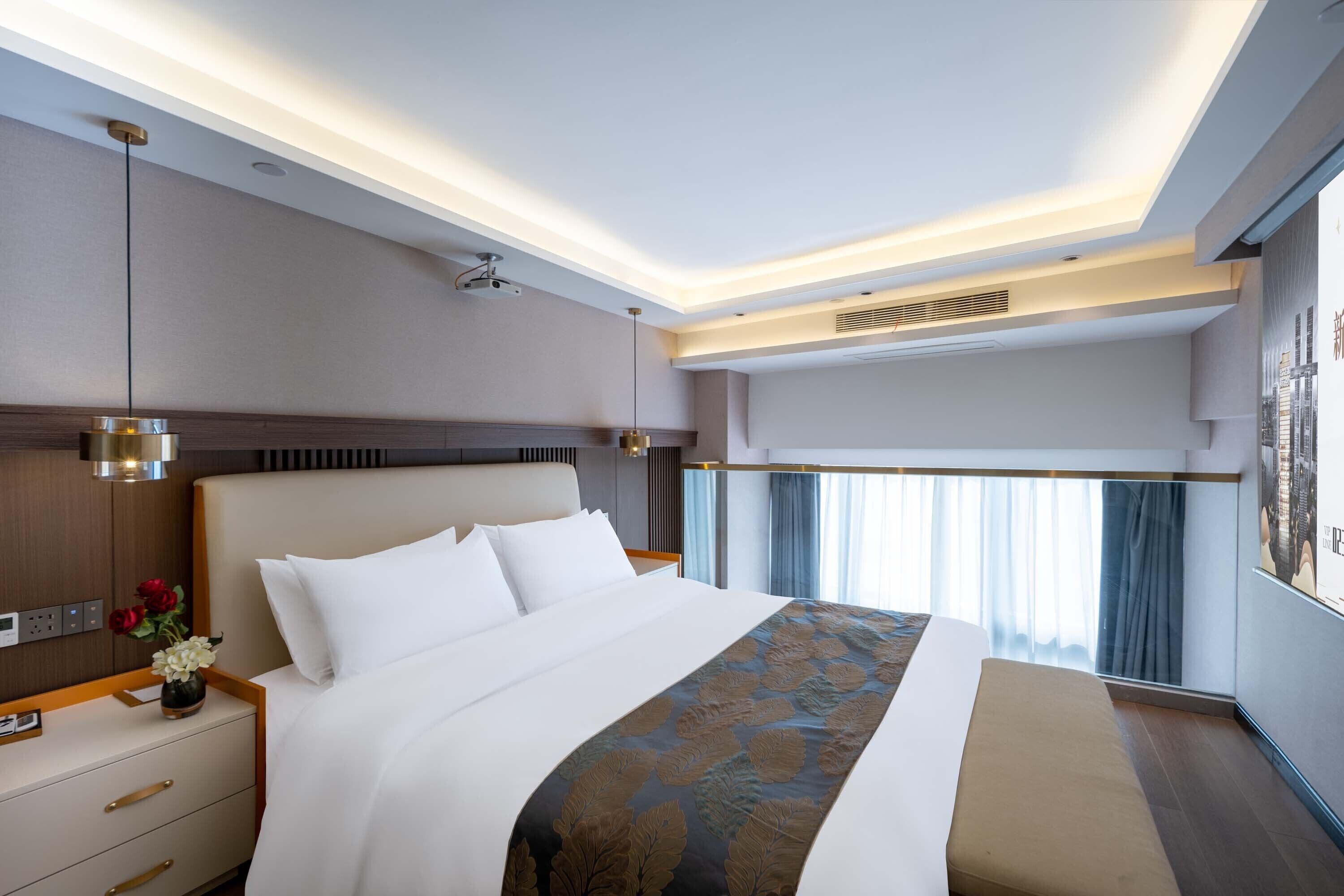 A modern hotel room with a large bed, a bedside table with a vase of flowers, a bench, and a large window with curtains.
