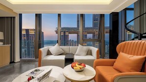 Una habitación de hotel moderna con un ventanal grande, un sofá, un sillón naranja y una mesita con fruteros.