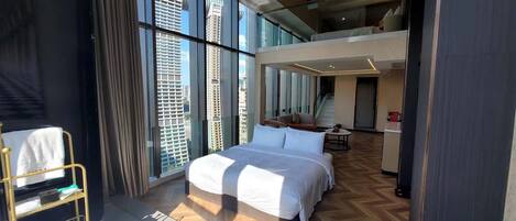 Una habitación de hotel moderna con una cama grande, un área de descanso con un sofá y sillones, una mesita y vistas a edificios altos a través de amplios ventanales.