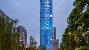 Un rascacielos moderno con fachada de vidrio reflectante, rodeado por un parque urbano bien iluminado que incluye una fuente y árboles.