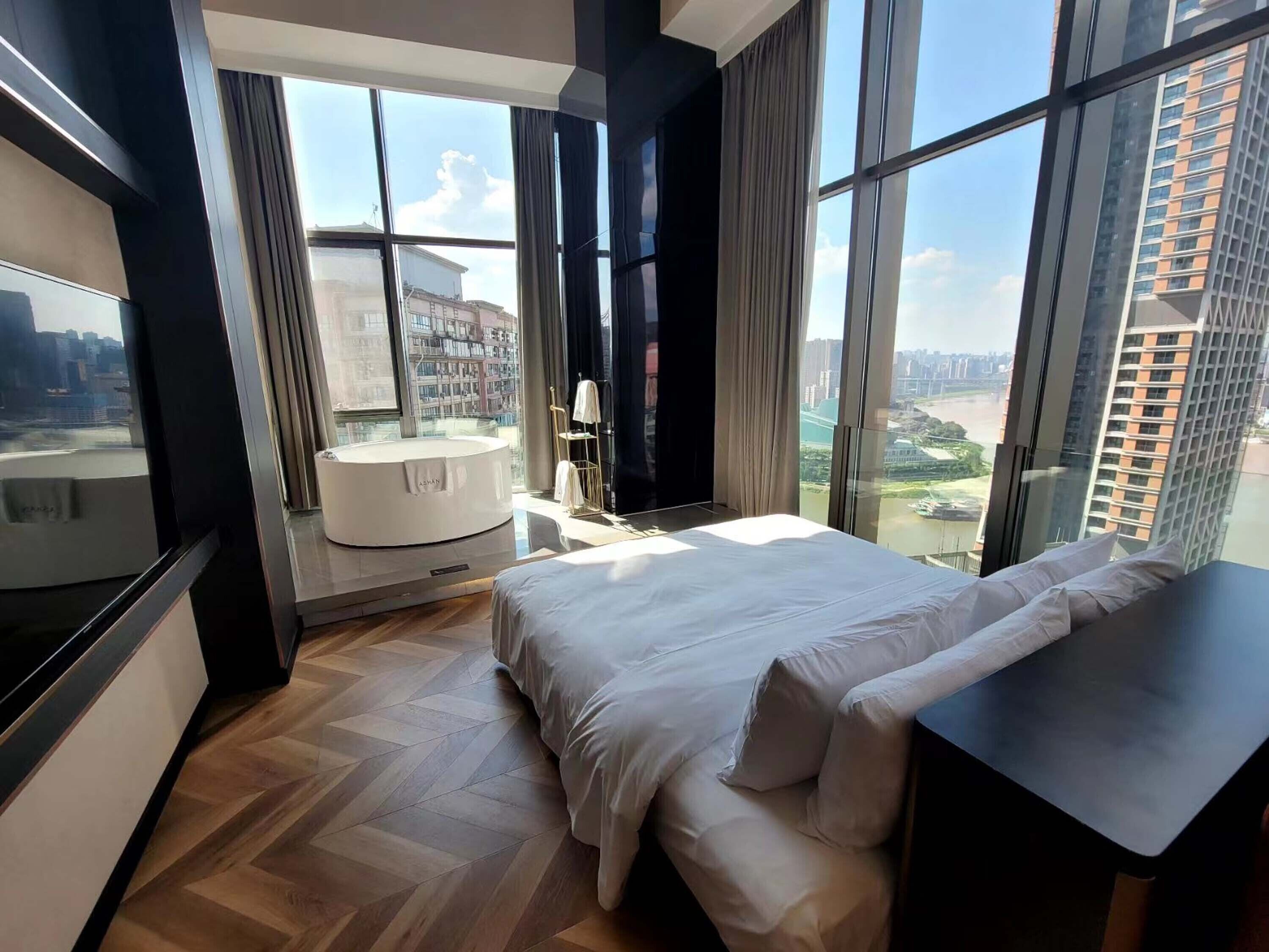 A modern hotel room with a large bed, a round bathtub, and a city view through large windows.