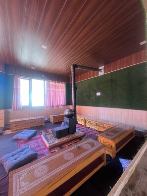 Une pièce aux murs et au plafond en bois, avec une grande fenêtre ornée de rideaux, un poêle à bois servant de chauffage central et des meubles traditionnels en bois.