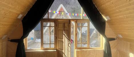 A wooden room with a sloped ceiling, a bed, a small table, and a view of a building outside.