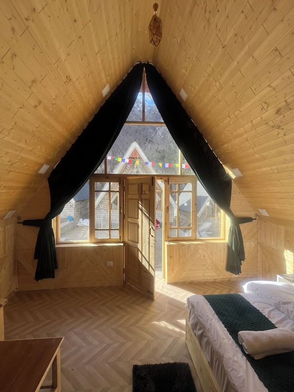 A wooden room with a sloped ceiling, a bed, a small table, and a view of a building outside.
