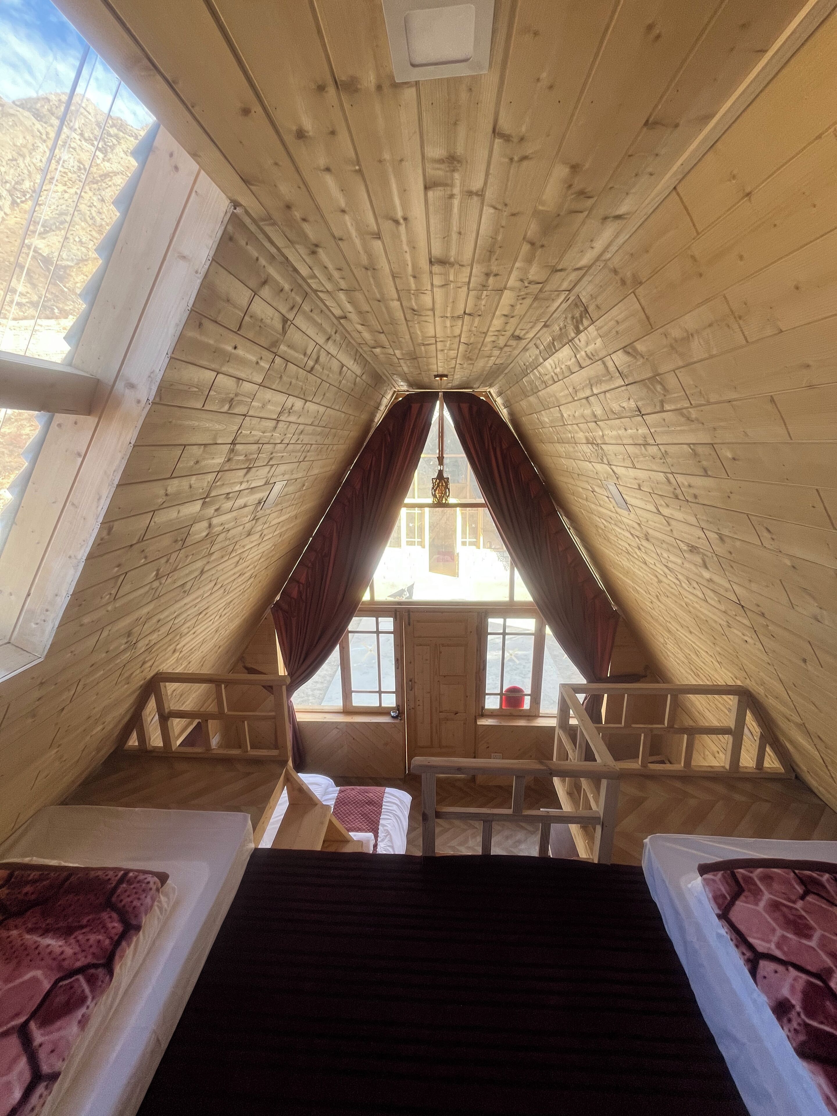 A high-ceilinged attic bedroom with a wooden sloped roof, a central window with curtains, and a bed with a patterned bedspread.
