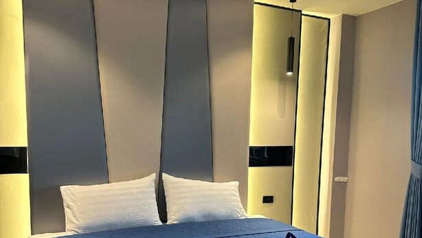 A modern hotel room with a large bed, a headboard with vertical panels, and a blue quilted bedspread. - Puttaya Glow (Pattaya)