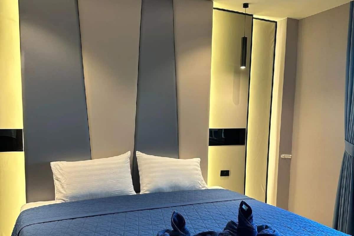 A modern hotel room with a large bed, a headboard with vertical panels, and a blue quilted bedspread.