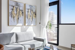 Ein modernes Wohnzimmer mit einem grauen Sofa, zwei abstrakten Gemälden, einem Glastisch und Blick auf einen Wolkenkratzer durch ein großes Fenster.