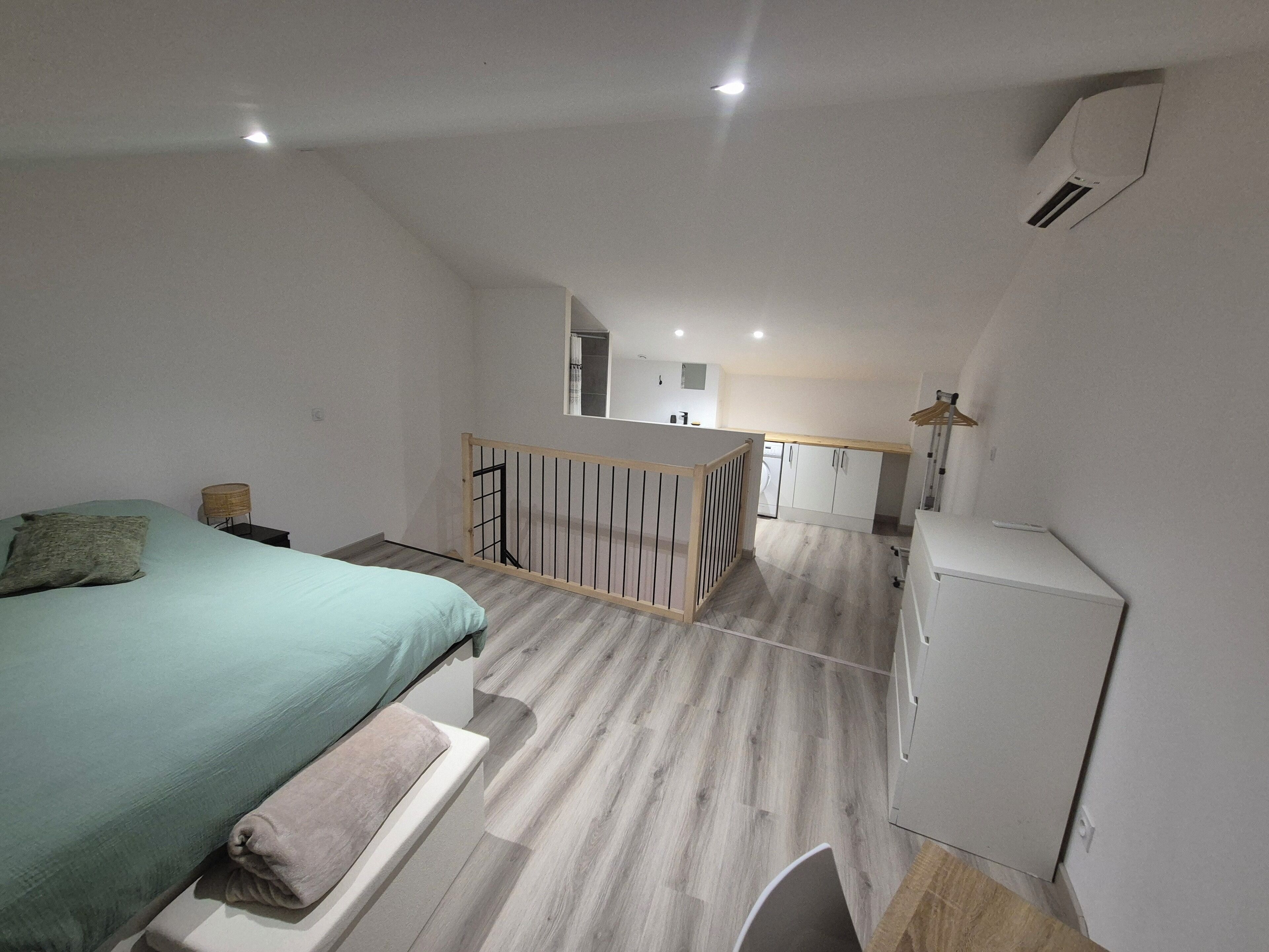 A bedroom with a bed, a small bedside table, a wooden railing, and a view of a kitchen and balcony.