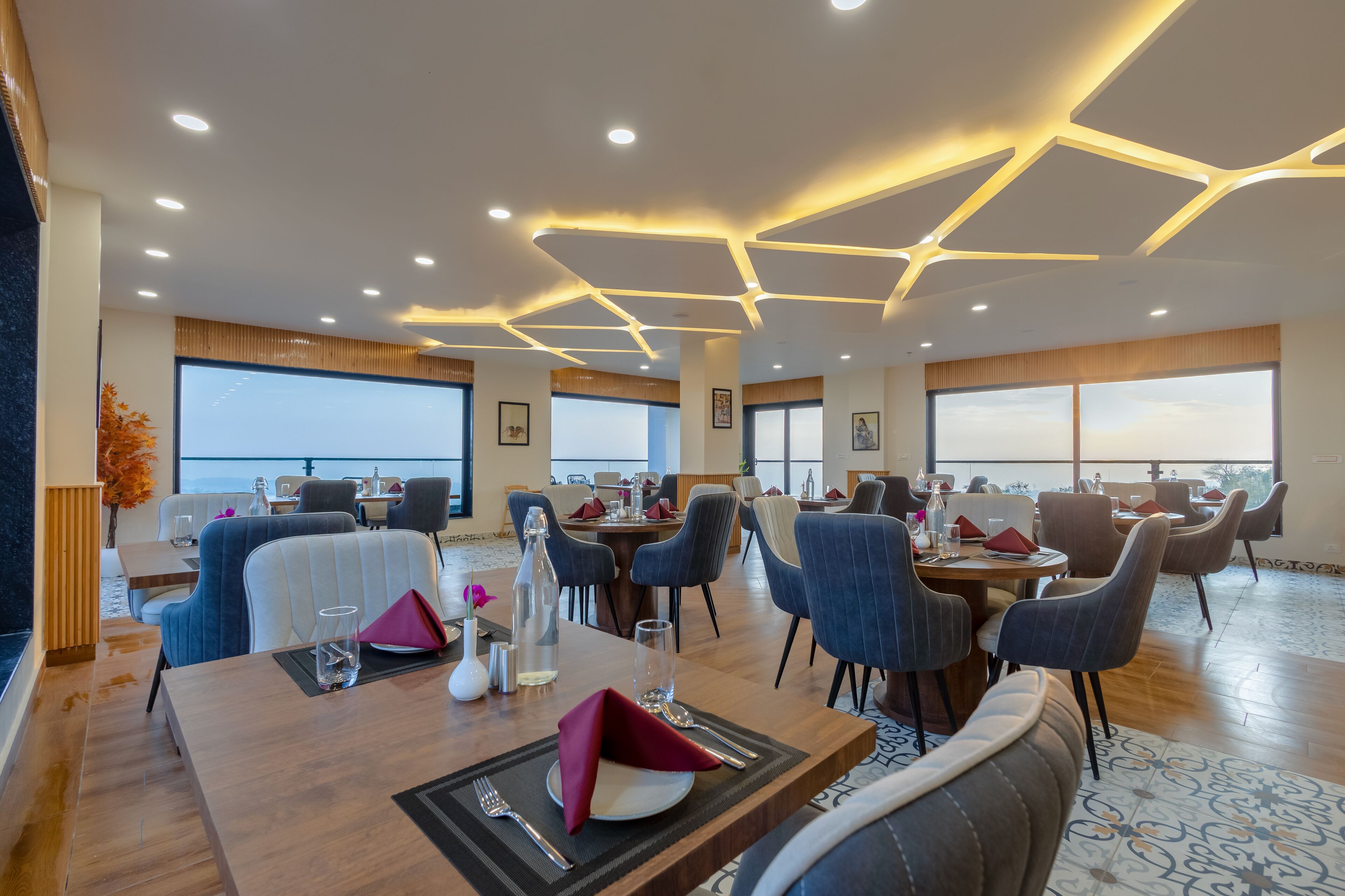 A modern dining area with tables set for service, large windows offering a view, and a geometric ceiling design.