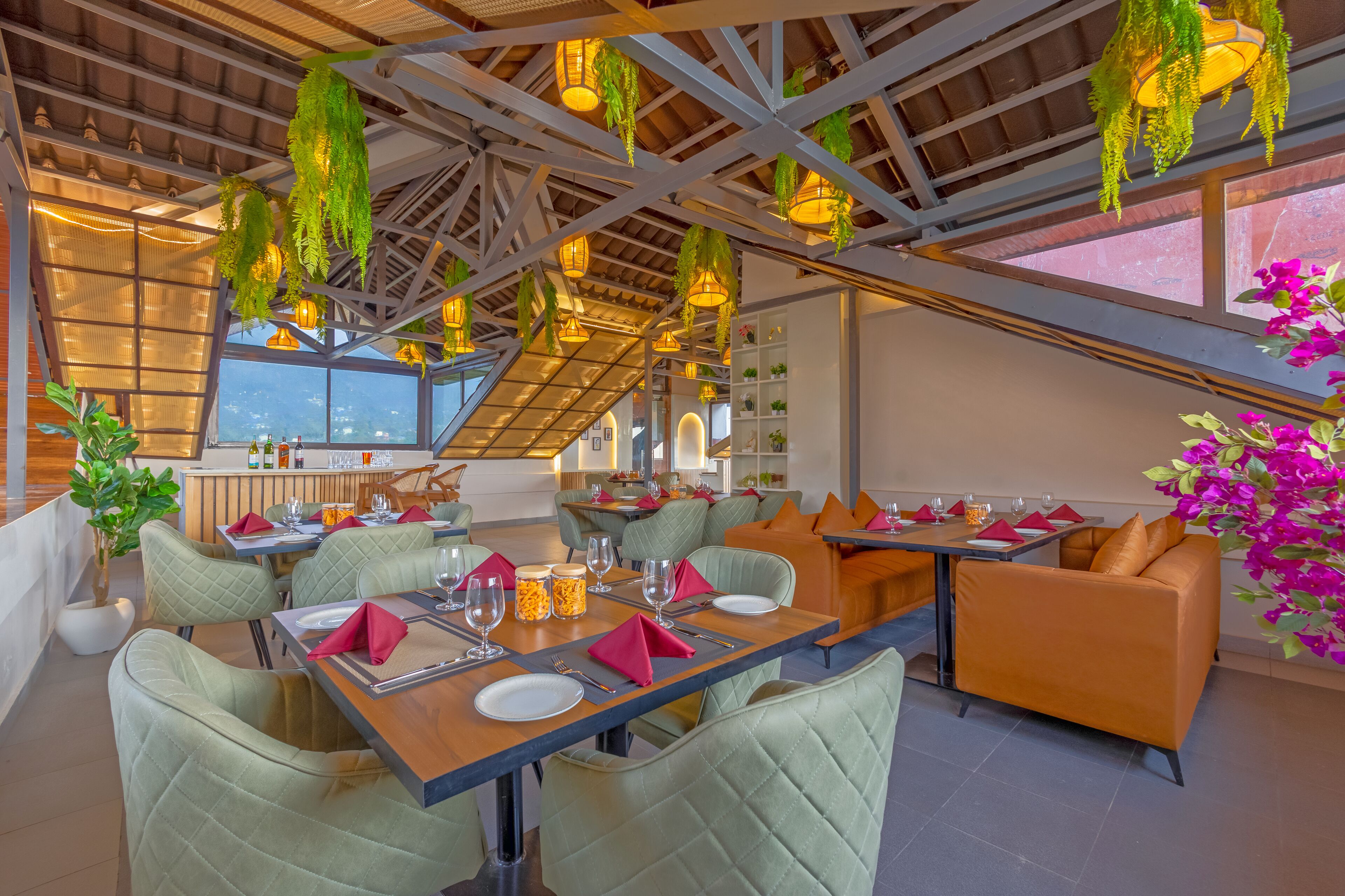 A restaurant with high ceilings, hanging plants, and a view of the city. Tables are set with red napkins and glassware.